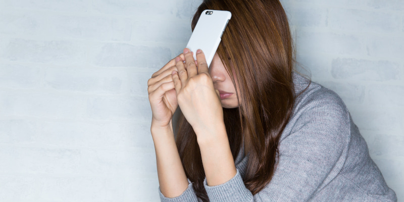 スマートフォンの下部を両手で持って額に当て、悩む女性の写真