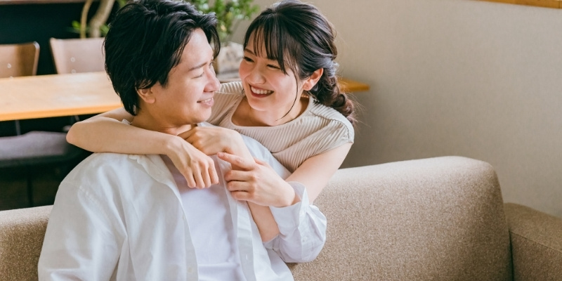 ソファに座る彼氏に背後から抱き着く彼女の写真