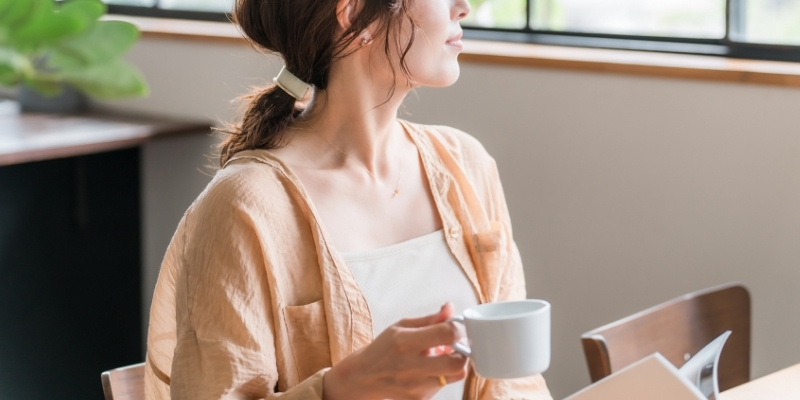 カフェでコーヒーを飲みながら、本を片手に外を眺めている女性の写真