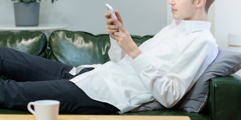 ソファに寝そべってスマホを操作する男性の写真