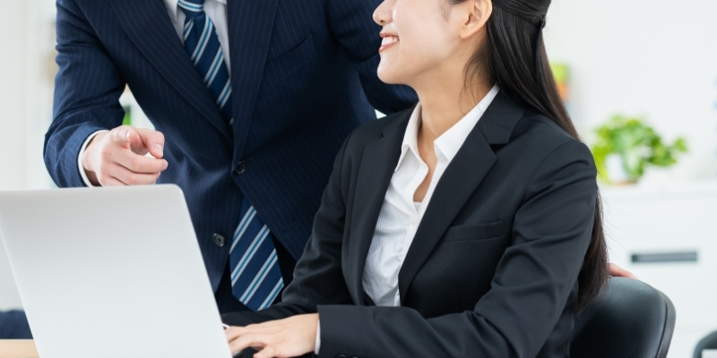 スーツ姿の男女の写真。男性がPCの前の女性に話しかけている
