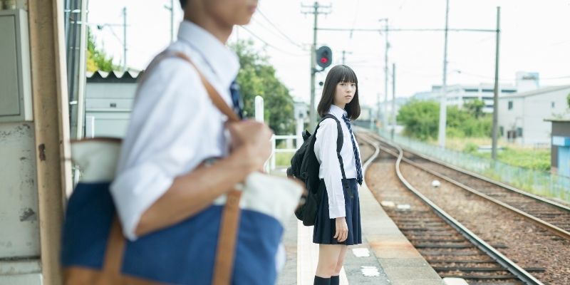 駅のホームで男子高校生を遠くから見つめている女子高生の写真
