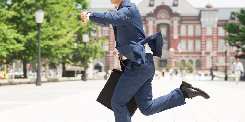 腕時計を見ながら東京駅の前を走るビジネスマンの写真