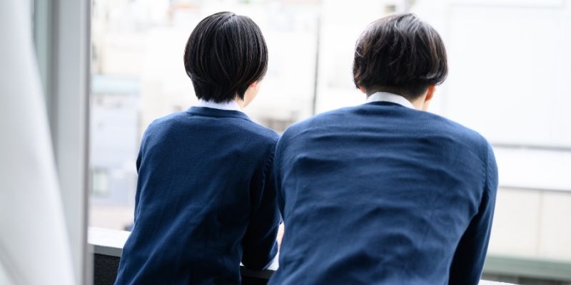 中高生くらいの学生カップルが窓に並んで喋っている後ろ姿の写真