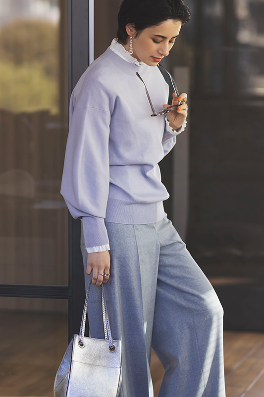 フリル白ブラウスにラベンダーニットを重ね着し、淡いグレーのパンツをはいたモデル写真
