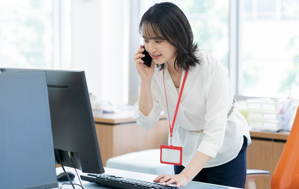 デスクトップパソコンを見ながらスマホで通話するビジネスウーマンの写真