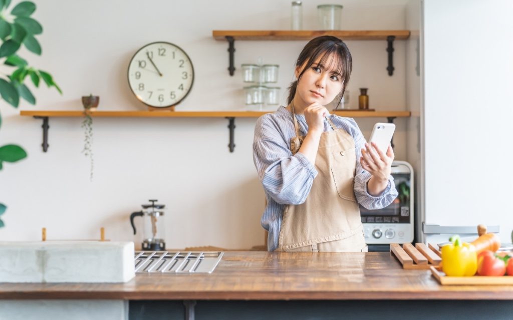 キッチンでスマホを片手に悩むエプロン姿の女性の写真