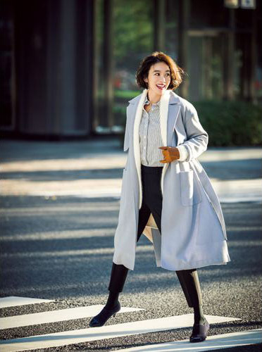 ストライプシャツに白のカーディガンを重ね着し、黒パンツをはいて淡い水色のロングコートをはおったモデル写真
