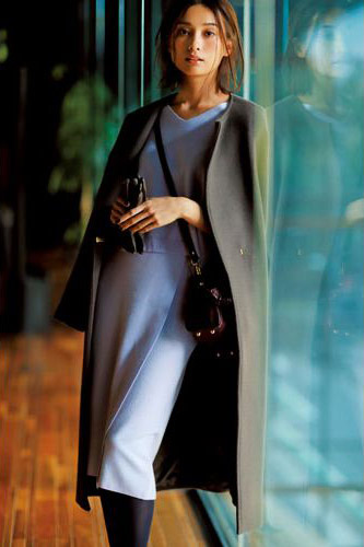 ラベンダー色のニットとスカートのセットアップに、茶色のノーカラーロングコートを肩がけしたモデル写真