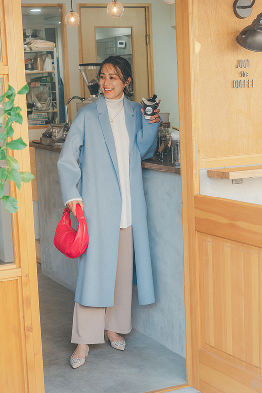 白ニットの上に白ブラウスを重ね着し、ベージュパンツをはいて水色のロングコートをはおったモデル写真