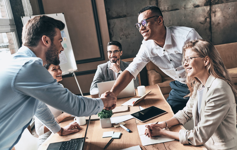 5人で会議中の風景。机を跨いで男性二人が握手している