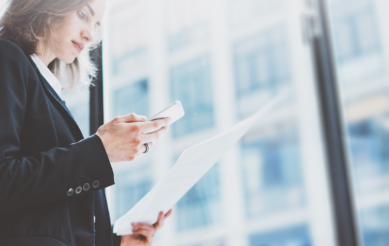 書類を片手に持ち、スマホを見るビジネスウーマンの写真