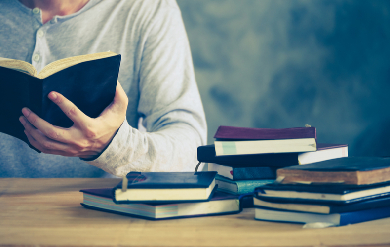 沢山の本が積まれたテーブルで本を読む男性の写真
