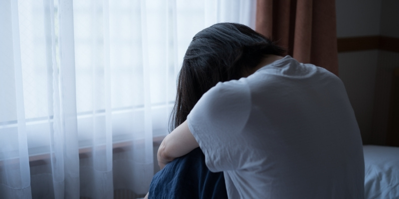 暗い部屋で膝を抱えて落ち込む女性の写真