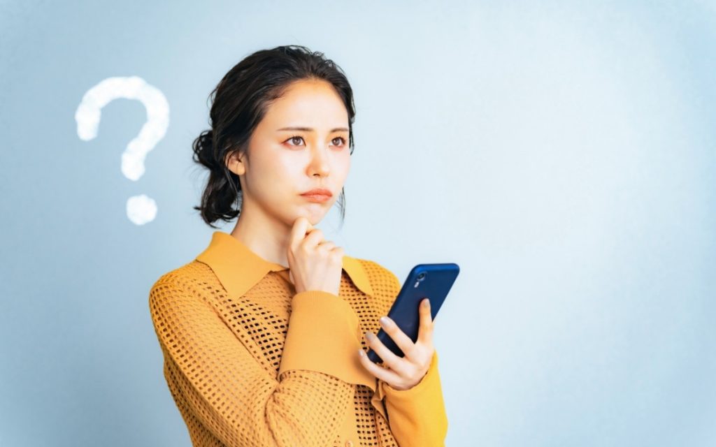 スマホを片手にはてなマークを出して考えている女性の写真