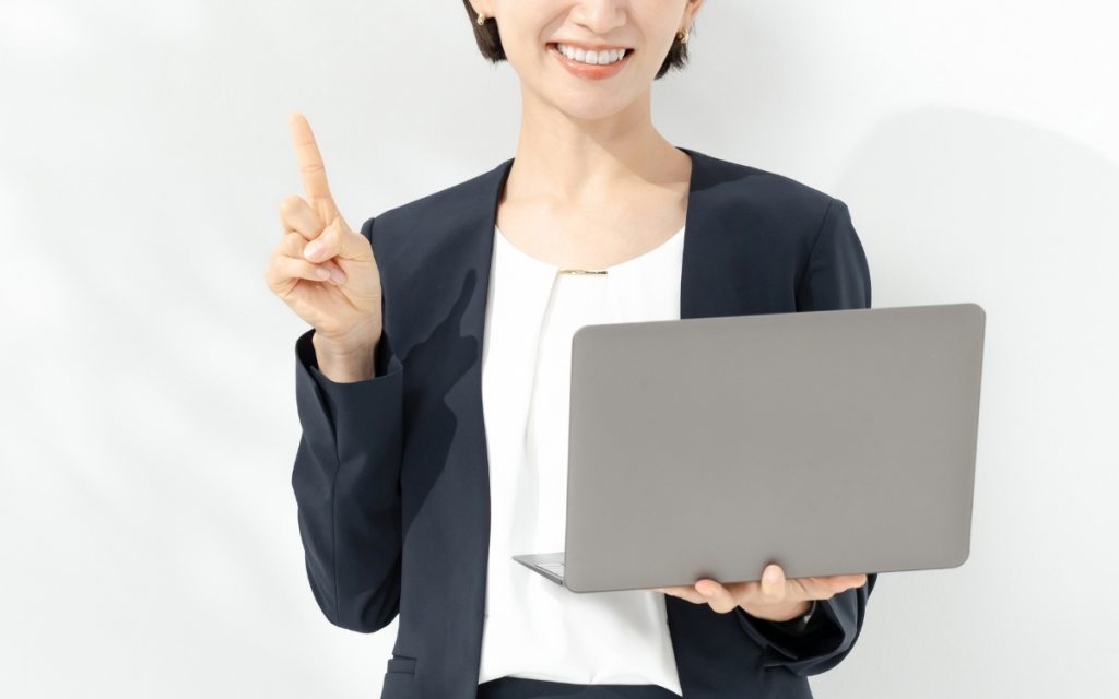 ノートパソコンを片手に人差し指を上に向ける女性の写真