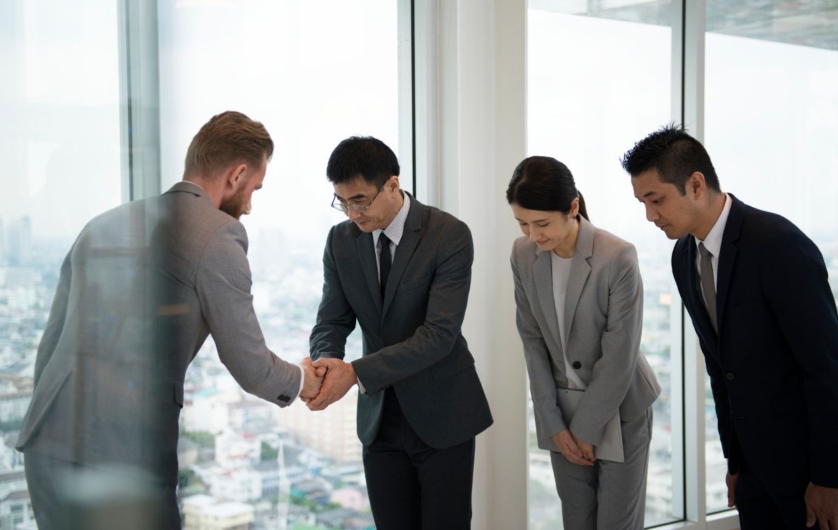 外国人男性と握手する日本人ビジネスマンの写真。日本人ビジネスマンの横では部下が二人お辞儀している