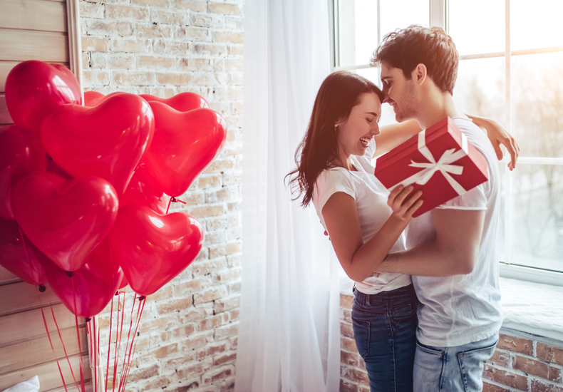 プレゼントを持った女性と抱き合う男性。部屋には赤いハートの風船が飾られている
