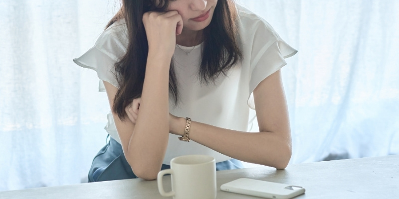 テーブルに置いたスマホを見ながら頬杖をついて考え込む女性の写真